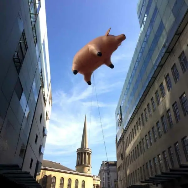 Colorful pig-shaped balloon hovering between modern and historic buildings in the city.