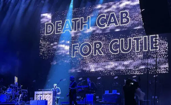 A vibrant stage featuring musicians with a big screen displaying "DEATH CAB FOR CUTE" during a live concert event.