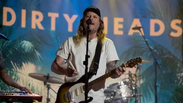 A musician with long hair and tattoos singing and playing guitar at a concert with a lively, tropical-themed backdrop.