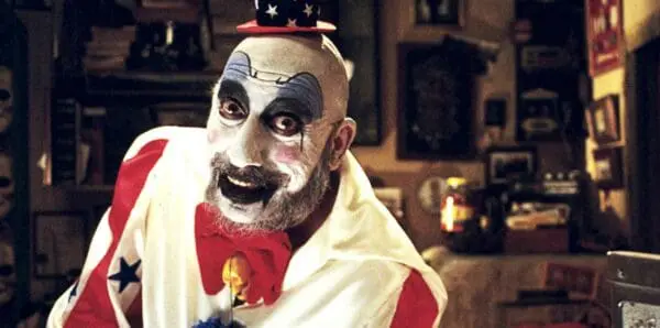 Clown in patriotic attire with face paint, wearing an Uncle Sam hat, in a cozy room.