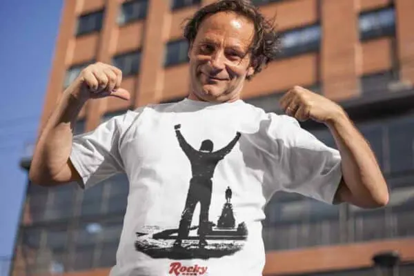 Man wearing a Rocky-themed T-shirt outdoors, flexing muscles in front of a building.