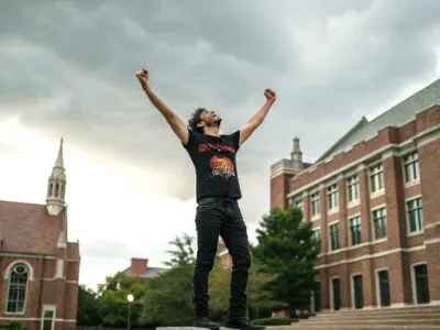 A person celebrating outdoors on a college campus with raised fists, symbolizing victory and empowerment, in front of historic brick buildings.