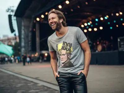 A smiling man at an outdoor music festival wearing a graphic T-shirt, enjoying the lively evening atmosphere with stage lights in the background.