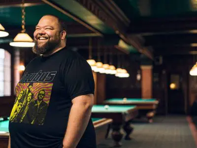 A smiling man wearing a graphic band t-shirt in a cozy billiards room with pool tables and warm lighting.