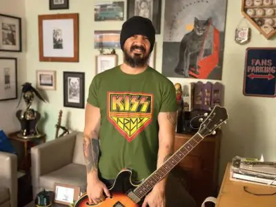 A man holding an electric guitar in a cozy, decorated room with rock-themed artwork and framed photos, showcasing stylish and casual fashion.
