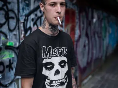 A young person with tattoos and a cigarette wearing a Misfits band t-shirt stands against a graffiti-covered wall, capturing edgy streetwear style.