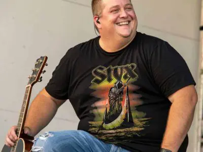 A happy man wearing a black Styx band t-shirt, playing an acoustic guitar on stage at a live concert.