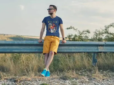 A young man relaxing outdoors during sunset, wearing a Weezer band t-shirt, yellow shorts, and blue sneakers by a metal guardrail.