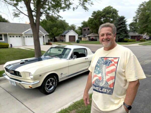 Vintage Shelby Mustang with man smiling, American flag-inspired Shelby T-shirt, residential street scene.