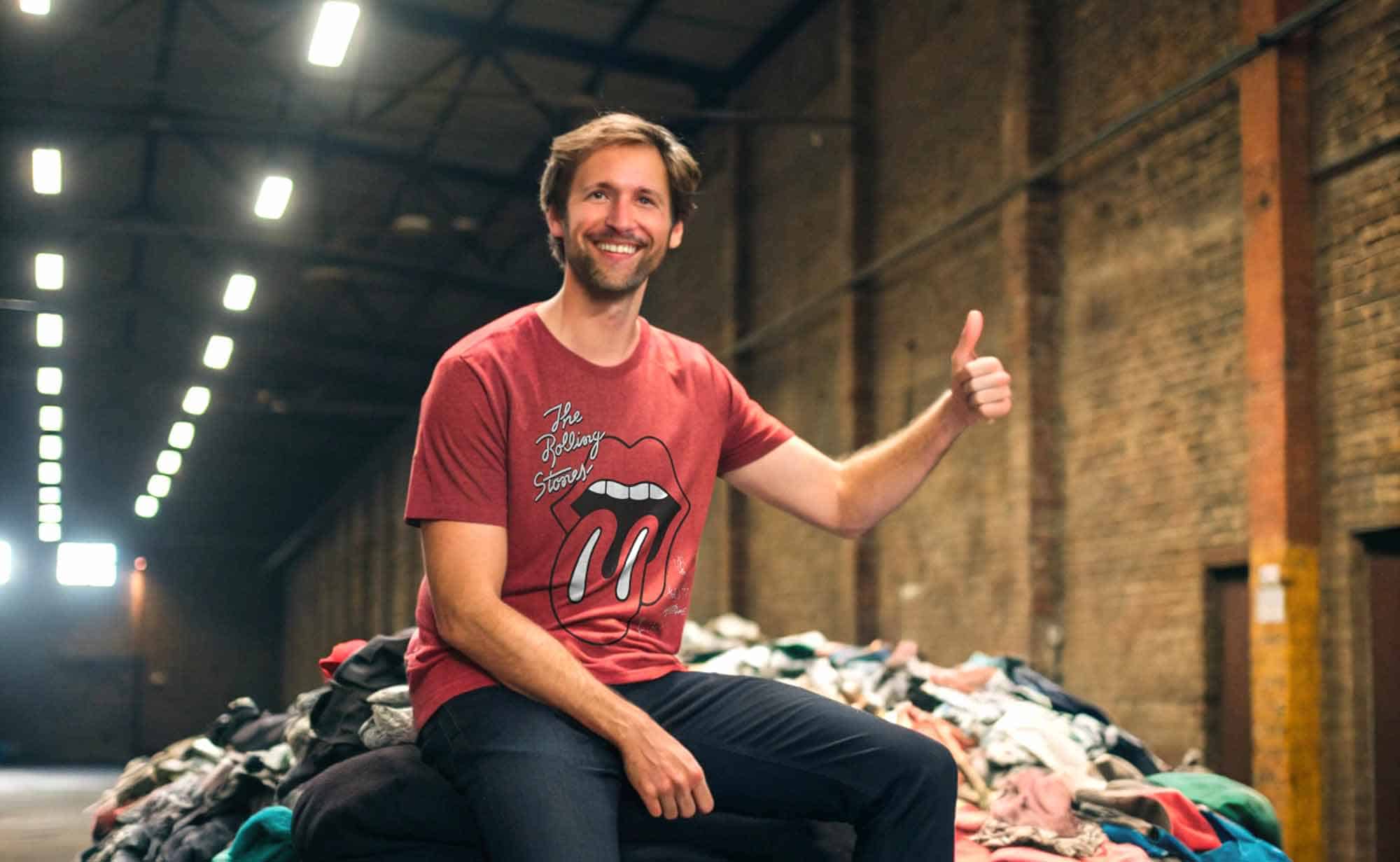 Alt: Man smiling and giving a thumbs-up while sitting on a mountain of clothing in an industrial space.