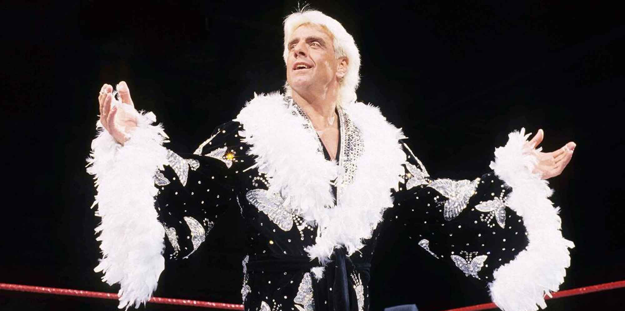 Iconic wrestling star in ornate robe with feathered cuffs, mid-performance on stage.