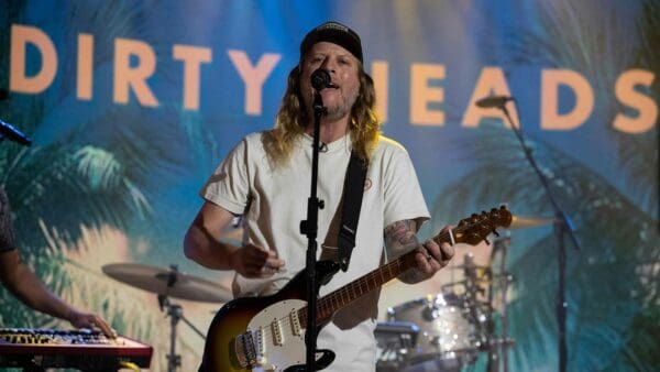 A musician with long hair and tattoos singing and playing guitar at a concert with a lively, tropical-themed backdrop.