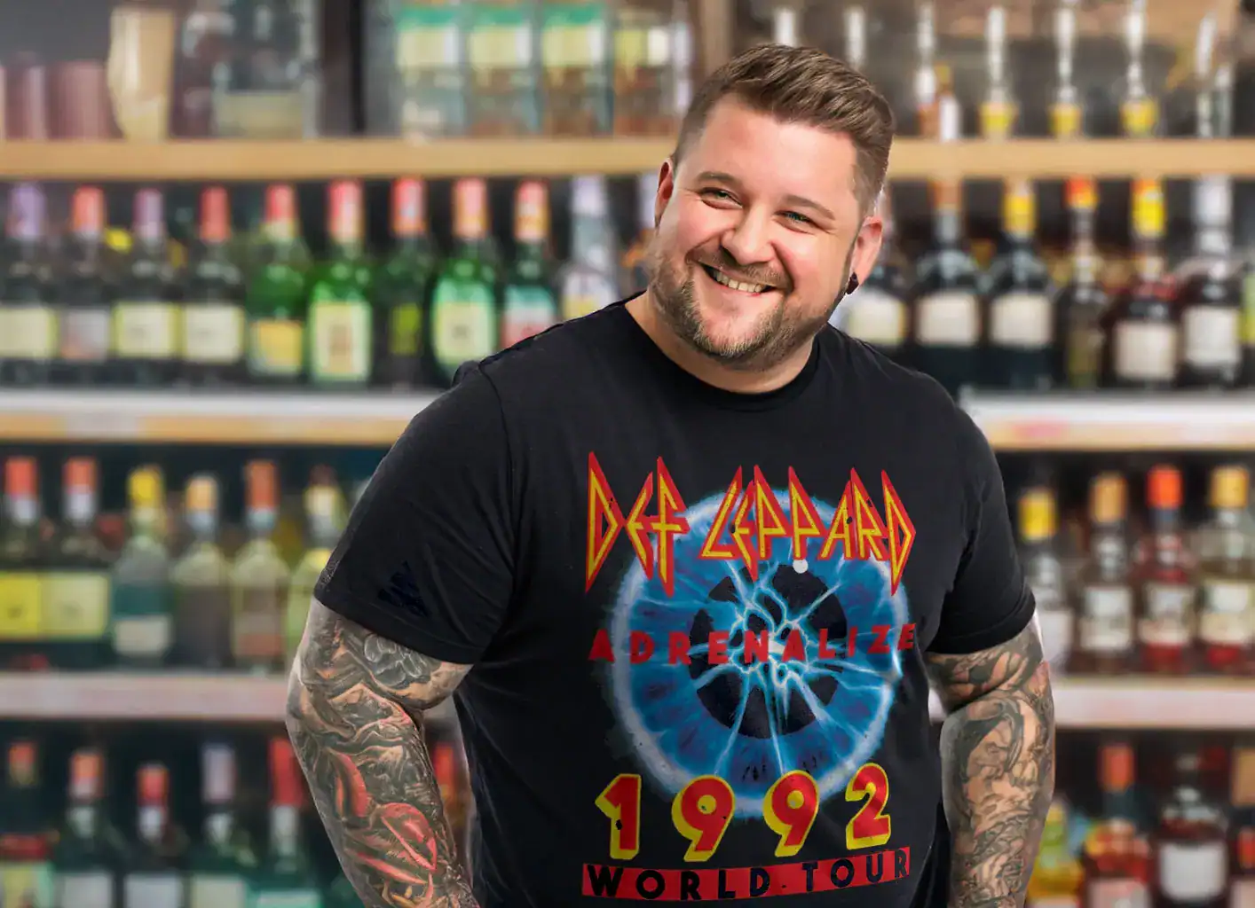 Young man smiling in a liquor store, wearing a Def Leppard Adrenalize 1992 Tour T-shirt, surrounded by shelves of alcohol bottles.