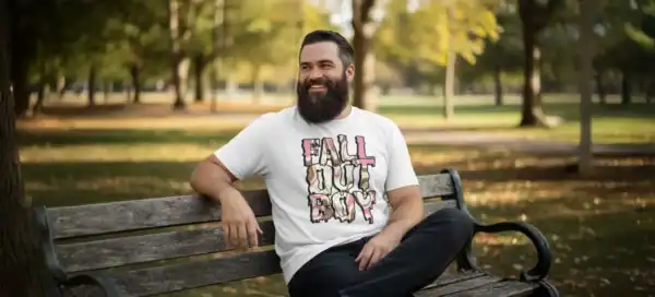 A man with a beard smiles while sitting on a park bench during fall, wearing a "Fall Out Boy" graphic T-shirt, surrounded by colorful autumn trees and a peaceful outdoor setting.