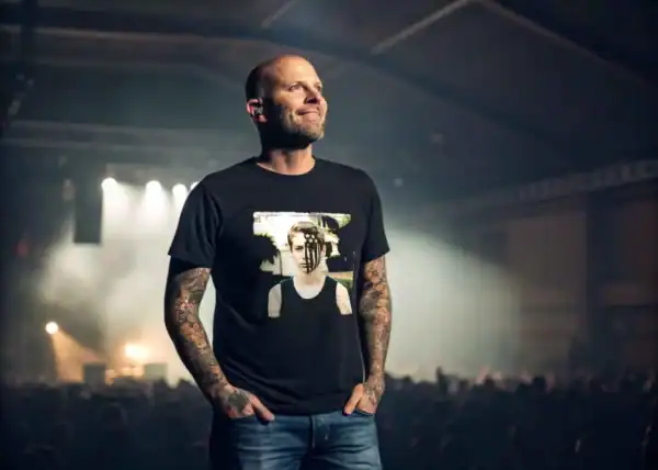 A tattooed man in a graphic t-shirt attending a live concert with stage lighting and an energetic atmosphere.