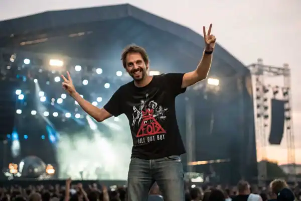 A cheerful man enjoying a live rock concert at an outdoor festival, wearing a Fall Out Boy band T-shirt amidst stage lights and a large crowd.