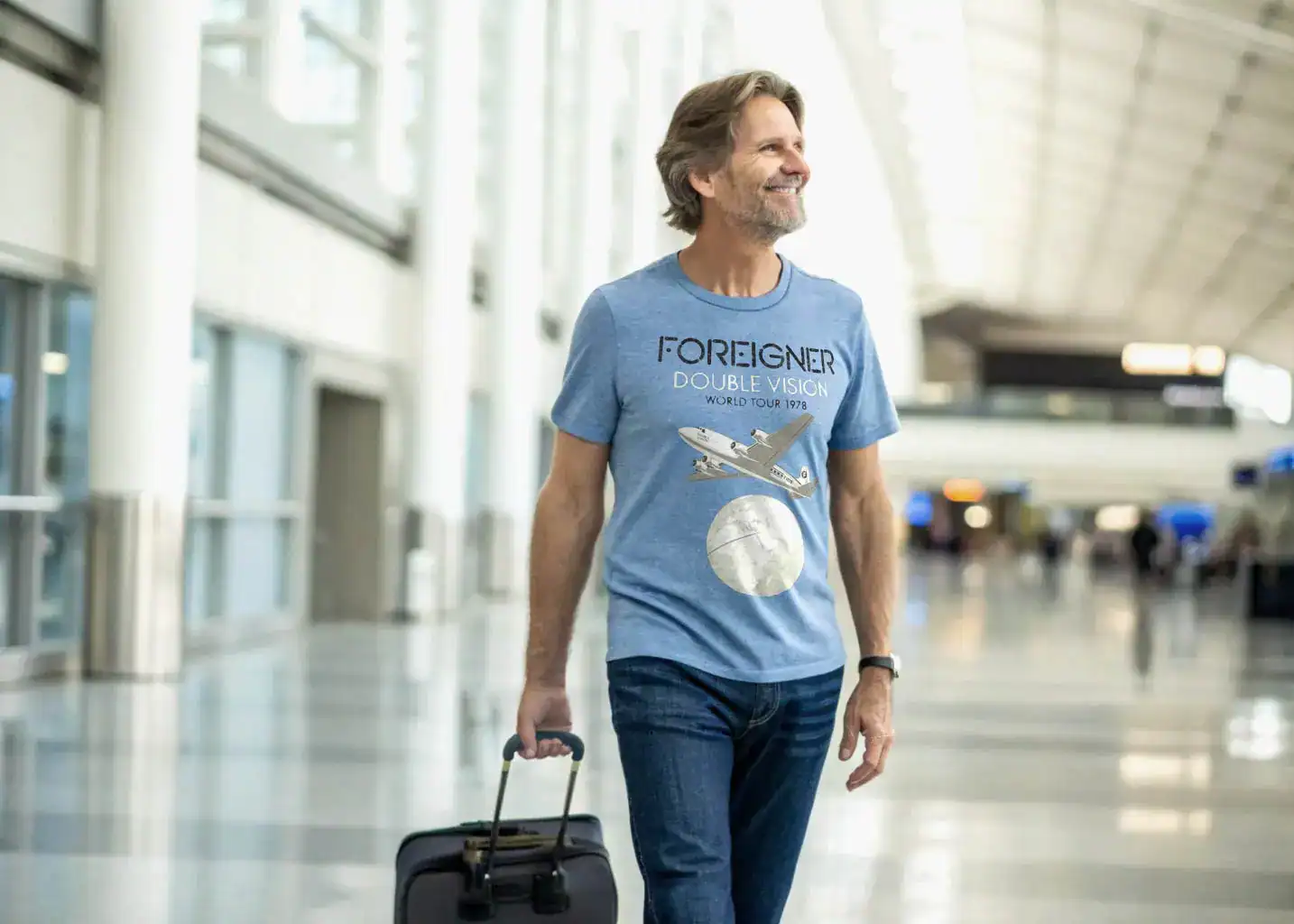 A middle-aged man walking through an airport wearing a Foreigner Double Vision World Tour 1978 Men’s T Shirt