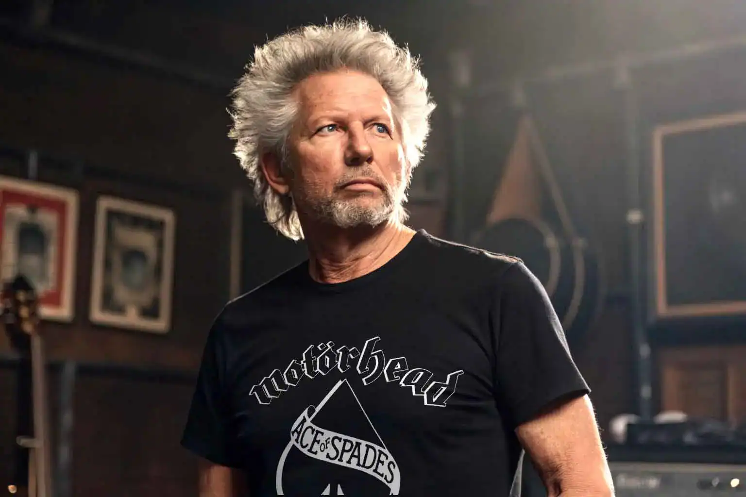 A stylish older man with white hair wearing a Motörhead T-shirt in a creative studio or music space.
