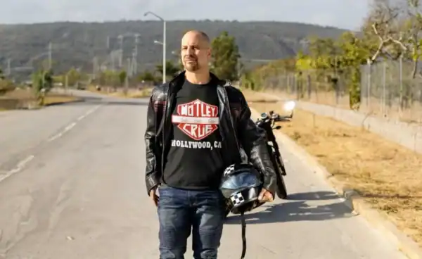 Man in a black leather jacket wearing a Mötley Crüe “Hollywood, CA” t-shirt stands on an open road holding a motorcycle helmet, with a motorcycle behind him.
