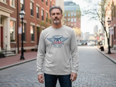 A stylish man wearing a graphic sweatshirt featuring a vintage-inspired design with wings and text, standing on a charming city street in natural light.