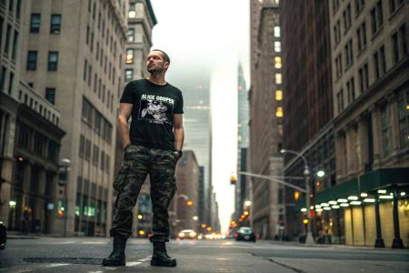 A man stands confidently in the middle of a city street during evening, wearing a black graphic t-shirt and camouflage pants, showcasing urban fashion style.