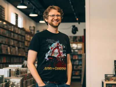 Alternate text: Man wearing Army of Darkness T-shirt in record store, smiling and browsing music.
