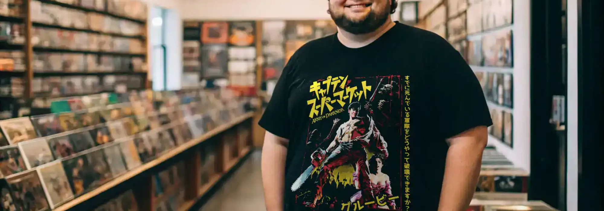 The man wears a Japanese anime-inspired t-shirt with a bold design, standing in a record store with shelves of vinyls behind him.