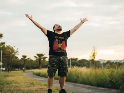 Man enjoying outdoor freedom with arms raised, dressed casually, in a scenic park at sunset.