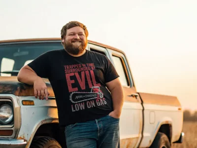 Vintage truck and man wearing humorous "Evil" T-shirt in rural setting at sunset.