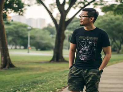 A man standing with hands in pockets in a lush park with trees and walking path, dressed casually with glasses.