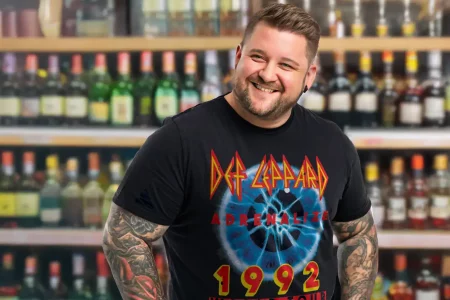 Young man smiling in a liquor store, wearing a Def Leppard Adrenalize 1992 Tour T-shirt, surrounded by shelves of alcohol bottles.