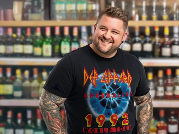Young man smiling in a liquor store, wearing a Def Leppard Adrenalize 1992 Tour T-shirt, surrounded by shelves of alcohol bottles.