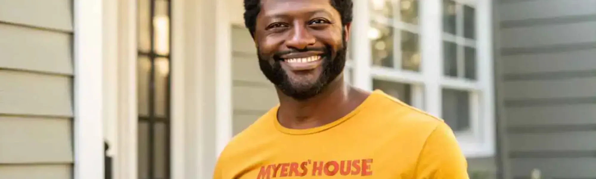 Happy man in yellow graphic tee standing outside in front of house, smiling for the camera.