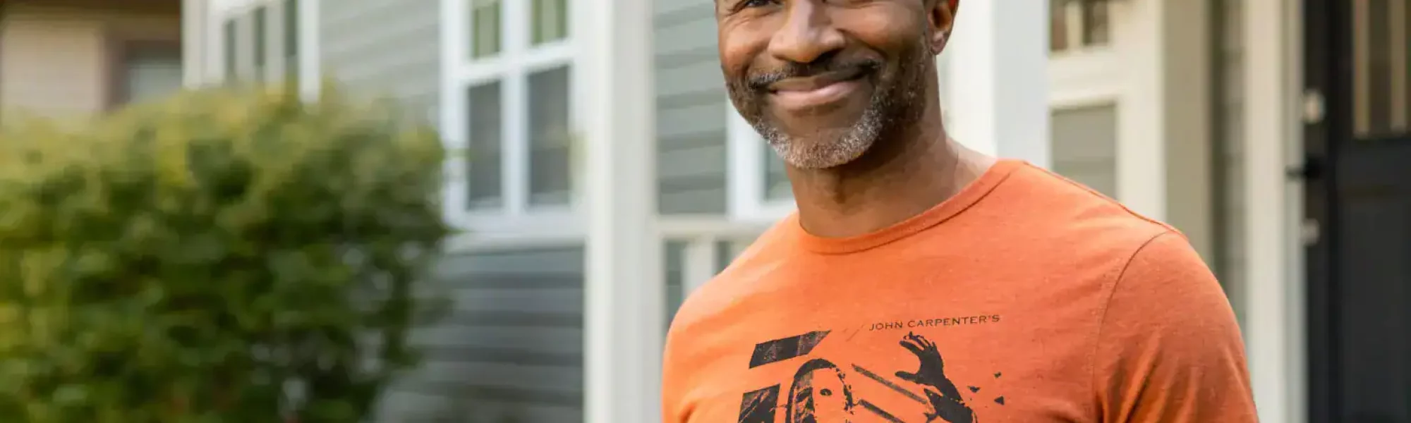 Smiling middle-aged man in Halloween T-shirt standing outside a house on a sunny day.