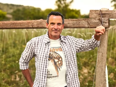 A man in casual checkered shirt and graphic T-shirt stands outdoors in a field, smiling confidently with a wooden fence in the background.