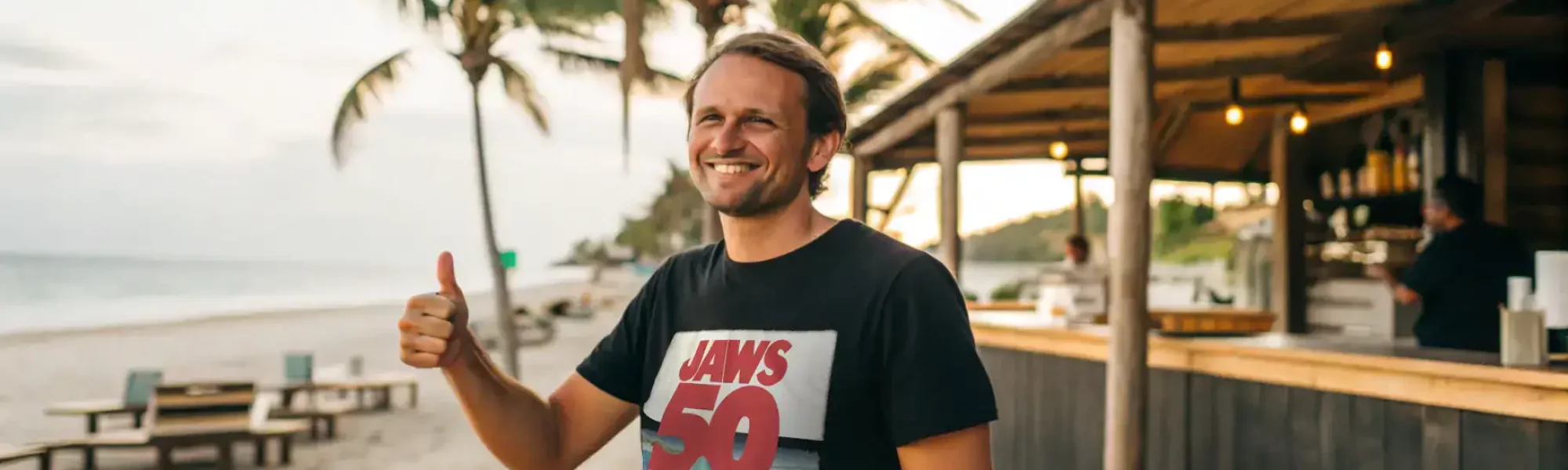 Vibrant beach scene with smiling man in Jaws t-shirt, palm trees, and lively beach bar, perfect for summer and lifestyle SEO.
