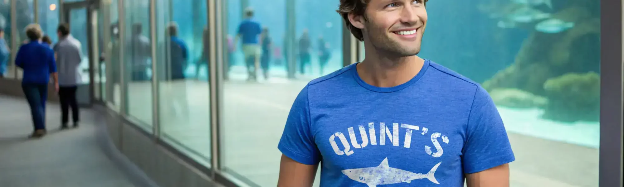 Colorful man in shark-themed T-shirt at aquarium with visitors in background.