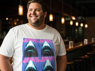 A smiling man stands in a warmly lit bar, wearing a Jaws Jaws graphic T-shirt, creating a casual and inviting atmosphere.