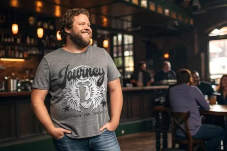 A man wearing a "Journey" graphic t-shirt, smiling in a lively, cozy bar setting with friends.