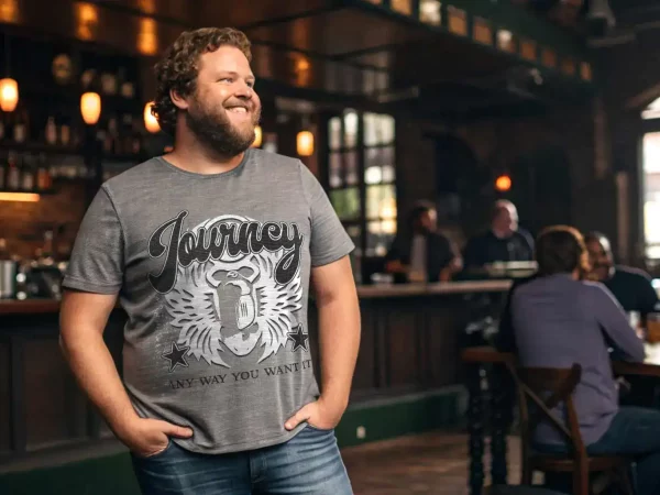 A man wearing a "Journey" graphic t-shirt, smiling in a lively, cozy bar setting with friends.