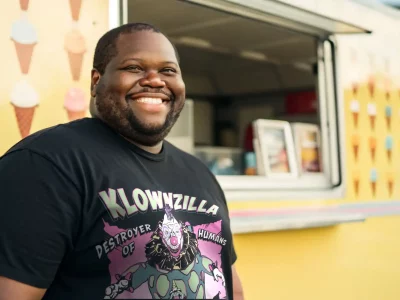 Happy man in clown-themed T-shirt standing near colorful ice cream truck at outdoor event.
