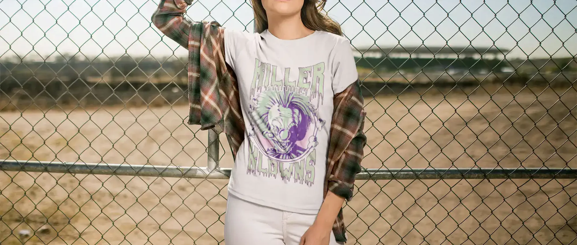 Stylish young woman wearing a "Killer Clowns" graphic t-shirt with a plaid shirt, standing by a chain-link fence on a deserted field.
