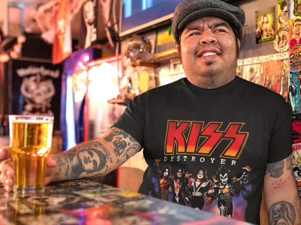 A man with tattoos wearing a KISS Destroyer T-shirt, sitting at a bar with a beer, surrounded by colorful stickers.