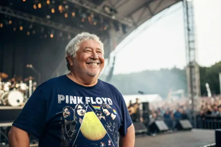 A smiling man at a rock concert wearing a Pink Floyd t-shirt, enjoying the outdoor music festival with a lively crowd and stage in the background.