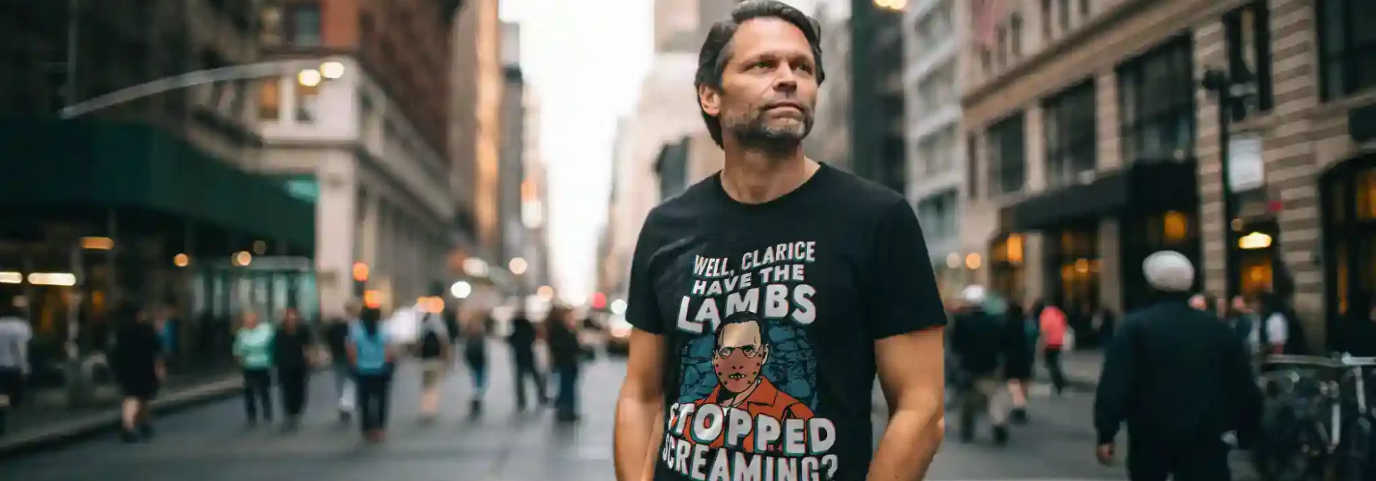 Man wearing a black T-shirt with humorous graphic and text standing in busy city street at dusk.
