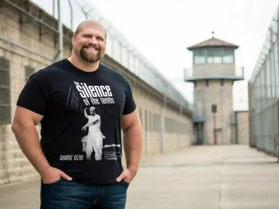 A man wearing a Silence of the Lambs t-shirt displaying Hannibal Lecter, standing outside a prison with guard towers, promoting horror-themed apparel.