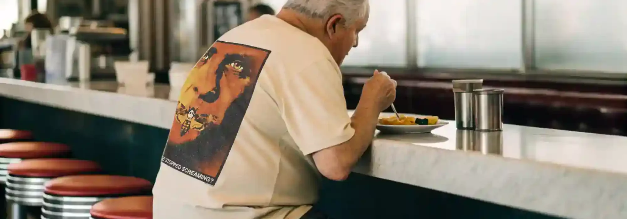 An elderly man with white hair eats alone at a retro diner counter, sitting on red stools, enjoying a meal on a sunny day.
