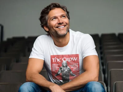 A cheerful middle-aged man with gray hair and beard, wearing a "The Crow" t-shirt, smiling in an empty auditorium.