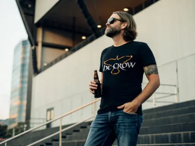 A man in sunglasses wearing a black SocieTees T-shirt with The Crow design and holding a beer, standing outdoors in an urban environment.