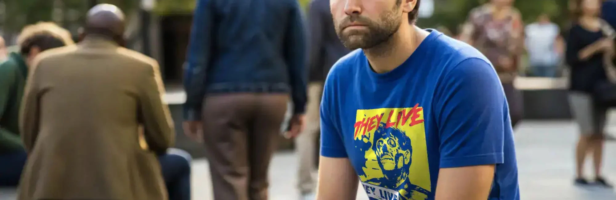 Portrait of a man with a serious expression wearing a colorful graphic T-shirt, sitting outdoors on a park bench with people in the background.